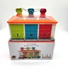 A toy garage with three doors, each a different color and with a different shape cutout. The garage has a transparent orange roof and three pull-tabs on top. It is shown on top of its packaging. Voir les images du produit