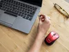 A person is plugging a USB dongle into a laptop while a red wireless mouse sits on a wooden desk next to a pair of glasses. Anteprima dell’immagine prodotto