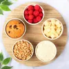 A top-down view of five small white bowls arranged in a circle on a wooden board. The bowls contain various food items: raspberries, cubed cheese, peanuts, a muffin, and a creamy dip. Produktbild-Vorschau