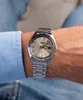 A person wearing a blue shirt and jeans, showing off a silver Seiko watch on their wrist. Produktbild-Vorschau