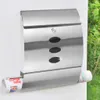 A stainless steel mailbox mounted on a white wall, with greenery in the background. A rolled-up newspaper is visible in the bottom slot. Produktbild-Vorschau