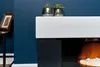 A close-up view of a modern electric fireplace with a white mantelpiece and a dark anthracite base. The fireplace has a realistic flame effect with white stones at the bottom. Two small potted plants in metallic pots sit on the mantelpiece. A framed pictur Náhled obrázku produktu