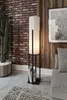 A floor lamp with a white cylindrical shade and a black metal frame, placed next to a brick wall and a cactus. Books are stored in the base of the lamp. Produktbild-Vorschau