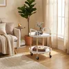 A round, two-tiered serving cart with wheels, styled in a Scandinavian design, is placed in a living room setting next to a sofa and a large potted plant. The cart is white with wooden legs and handles. A book and a mug are on the top shelf, and a book is  Anteprima dell’immagine prodotto