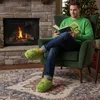 A man is sitting in a chair by a fireplace, reading a book and wearing Grinch-themed slippers and a sweater. Produktbild-Vorschau
