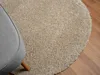 A close-up view of a round, beige shaggy rug with a portion of a gray armchair visible on the left, placed on a wooden floor. Náhľad obrázku produktu