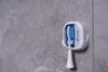 A white toothbrush sterilizer with a blue toothbrush inside, mounted on a gray tiled wall. Anteprima dell’immagine prodotto