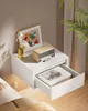A white floating nightstand is mounted on a wall next to a bed. It holds a small wooden radio, a framed colorful painting, and a vase with flowers. The nightstand has an open drawer revealing books and glasses. Náhled obrázku produktu