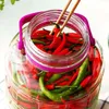 A close-up shot of a large glass jar filled with red and green chili peppers, with chopsticks holding a few peppers near the opening. Produktbild-Vorschau