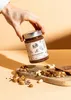 A hand holding a jar of spread over a wooden board with toast and nuts. Produktbild-Vorschau