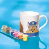 A white mug with Paw Patrol characters printed on it, along with small pots of paint and a paintbrush, set against a blue background. Produktbild-Vorschau
