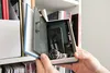 Hands holding an open book safe with a watch, phone, and wallet inside, on a bookshelf. Produktbild-Vorschau