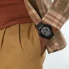 A close-up shot of a person wearing a digital watch on their wrist, with part of their plaid shirt and tan pants visible. Náhled obrázku produktu