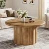 A round wooden coffee table with a fluted base is placed in a living room setting, next to a sofa and in front of a window with curtains. Produktbild-Vorschau