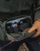 A person wearing gloves and a green jacket is opening a green hip pack, revealing its contents. Produktbild-Vorschau