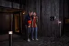 A couple stands outside a modern wooden house at night, illuminated by outdoor lighting. The man holds a red ball. Produktbild-Vorschau