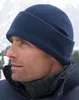 A man wearing a blue fleece hat and a dark blue jacket in a snowy mountain setting. Produktbild-Vorschau