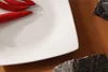 A close-up view of a white square plate with red chili peppers on it, placed on a brown surface with two dark stones. Náhled obrázku produktu