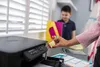 A person is refilling the ink tank of a printer while a child stands in the background holding school supplies. Podgląd zdjęcia produktu