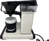 A white and black coffee maker with a white mug underneath the filter. Produktbild-Vorschau