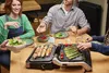 A group of people are gathered around a table, enjoying a meal cooked on a grill. Produktbild-Vorschau