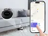 A black smart geo tag is shown on the floor near a sofa, with a smartphone displaying its location on a map. Náhled obrázku produktu