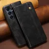 A black leather phone case with a gold snap closure is shown on a brown and tan textured background. Produktbild-Vorschau