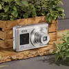 A silver digital camera is placed on a wooden surface, with a wooden crate and green plants in the background. Produktbild-Vorschau