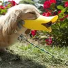 A dog wearing a yellow duck-shaped muzzle is in a grassy outdoor setting with red flowers in the background. Produktbild-Vorschau