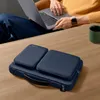 A person is using a laptop while a navy blue laptop bag with accessories sits on a wooden table. Produktbild-Vorschau