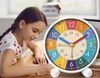 A young girl is studying at a desk next to a colorful alarm clock. Produktbild-Vorschau