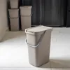 A taupe plastic trash can with a lid and handle sits on a white tiled floor in front of a dark appliance. Produktbild-Vorschau