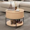 A round coffee table with two shelves is placed on a carpet in front of a sofa. The table has decorative vertical slats on its side and is adorned with books, vases, and a cup of coffee. Produktbild-Vorschau
