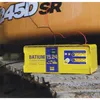 A yellow battery charger is placed on a rough, metallic surface, with part of a yellow vehicle in the background. Red and black cables from the charger are connected to the vehicle. Anteprima dell’immagine prodotto