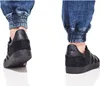 A close-up view of two feet wearing black Adidas Gazelle sneakers and cuffed denim jeans against a white background. Voir les images du produit