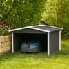 A robotic lawnmower is parked inside a small, dark gray shed with a white trim and a dark gray roof. The shed is situated on a green lawn, with a stone wall and pink flowers to the left, and trees and foliage in the background. Produktbild-Vorschau