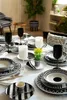 A dining table is set with a black and white patterned dinnerware set, complete with black wine glasses and white candles. Produktbild-Vorschau