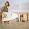 A woman holding a surfboard on a beach with a water bottle, a food container, and a straw bag on the sand. Náhled obrázku produktu