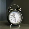 A black analog retro alarm clock with a white face is shown on a dark surface against a dark background. Náhled obrázku produktu