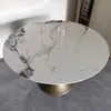 A round dining table with a marble-like ceramic top and a gold pedestal base is shown in a room. Produktbild-Vorschau