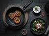 A top-down view of a frying pan with three patties, a plate with a patty and salad, and a bowl of dip, all arranged on a dark wooden surface with spices. Produktbild-Vorschau