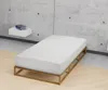 A minimalist bedroom scene with a concrete wall and a wooden floor. A white mattress on a wooden bed frame is the central focus, with a small white shelf holding two books on the wall. Produktbild-Vorschau