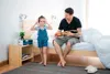 A father and daughter are in a bedroom. The father is sitting on the bed playing a ukulele, and the daughter is standing next to him wearing headphones. Podgląd zdjęcia produktu