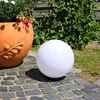 A white spherical lamp sits on a cobblestone path next to a terracotta planter filled with plants and flowers. A wooden fence and greenery form the background. Produktbild-Vorschau