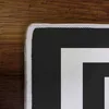 A close-up view of the corner of a black rug with a white border, placed on a wooden surface. Produktbild-Vorschau
