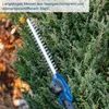 A close-up of a blue and silver hedge trimmer with a long blade, cutting into a green hedge. Produktbild-Vorschau