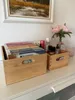 Two wooden storage boxes are on a white surface, one filled with books and the other with cosmetics. A painting hangs on the wall behind them, and a bouquet of pink flowers is in a vase. Produktbild-Vorschau