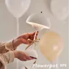 A hand holding a white mushroom-shaped lamp with a ribbon tied around its stem, with blurred balloons in the background. Produktbild-Vorschau