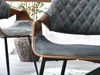 Close-up view of two modern dining chairs with upholstered seats and backs, featuring a diamond-stitched pattern on the backrest and curved wooden armrests. The chairs have black metal legs and are placed in a home environment with a woven basket and plant Náhled obrázku produktu