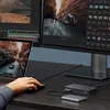 A person's hand is using a mouse connected to a laptop, which is part of a multi-monitor setup with a docking station and external hard drive. Produktbild-Vorschau